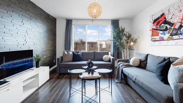 Een moderne woonkamer met een bakstenen muur, een groot raam, een grijze bank, een salontafel en een televisie op een witte console.