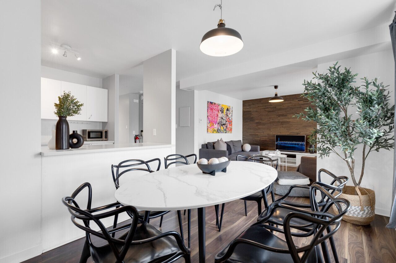 A modern kitchen with white cabinets, a dining area with a marble table, black chairs, and a large potted plant.