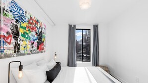Quarto moderno com uma pintura abstrata grande, cama com roupa de cama branca, luminária de cabeceira e vista da cidade pela janela.