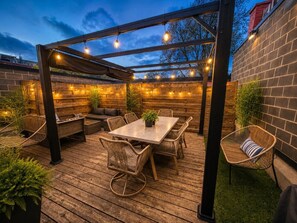 Un patio al aire libre con mesa y sillas de madera, una pérgola con luces colgantes y un muro de ladrillo.
