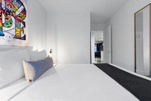 A modern hotel room with a large bed, a colorful abstract painting on the wall, and a view into the hallway.