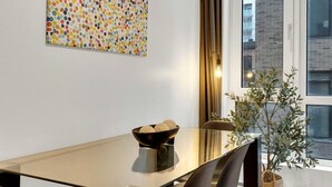 Et moderne spiseområde med et glasbord, en skål med runde genstande, en potteplante og et farverigt abstrakt maleri på væggen.