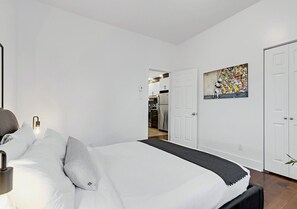 Un dormitorio con cama, una mesita de noche, una lámpara de pared y una puerta que da a la cocina.