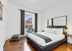 A bedroom with a large bed, a bedside table, a window with curtains, and a view of the outside.