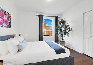 A bedroom with a bed, a window with curtains, a potted plant, and a door.