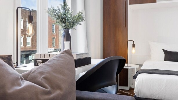 A modern hotel room with a large window, a desk with a vase of flowers, a bed with white bedding, and a nightstand with a lamp. - LiV MTL MTL Plateau Urban Suites Near Downtown Montreal (Montréal)