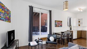 A modern living room with a sofa, a dining table, and a TV. There is a window with curtains, a kitchen area, and artwork on the walls. - LiV MTL MTL Plateau Urban Suites Near Downtown Montreal (Montréal)