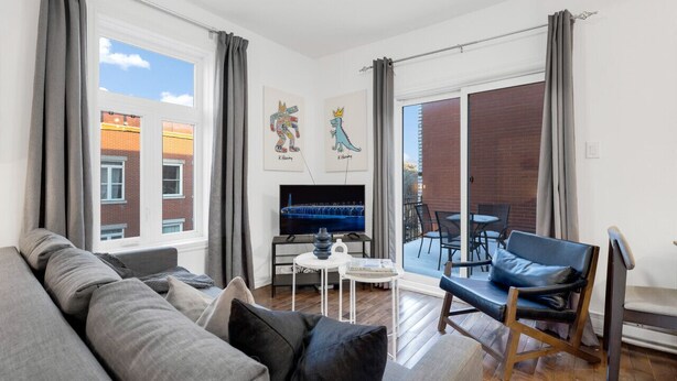 Un salon moderne avec un canapé gris, un fauteuil bleu, une petite table avec une lampe et un téléviseur. Il y a deux fenêtres avec des rideaux, un balcon avec une table et des chaises, et des œuvres d’art accrochées au mur.
