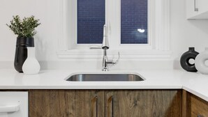 Une cuisine moderne avec un plan de travail blanc, un évier en acier inoxydable, des armoires en bois et une fenêtre dotée de vitres teintées en bleu.
