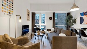 A modern living room with a sofa, chairs, a TV, a dining area, and a large window with curtains.