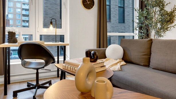 A modern living room with a sofa, a chair, a table with decorative items, a clock, and a view of buildings through large windows.