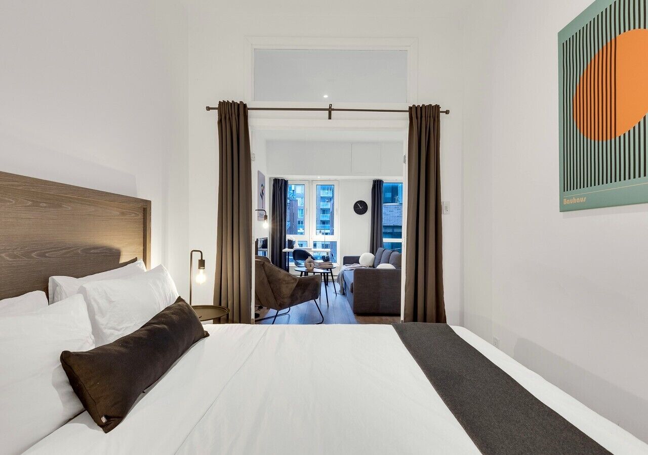 Habitación de hotel con una cama grande, vista de la ciudad a través de una ventana y un área de comedor al fondo.
