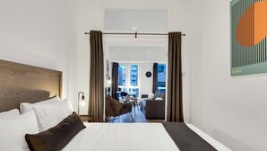 A hotel room with a large bed, a view of the city through a window, and a dining area visible in the background.