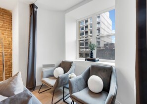 Un salon moderne avec un mur en briques, deux fauteuils gris dotés de coussins blancs, un canapé gris et une grande fenêtre donnant sur un immeuble.