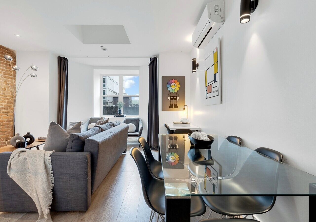 A modern living room with a glass dining table, grey sofa, and a brick wall.