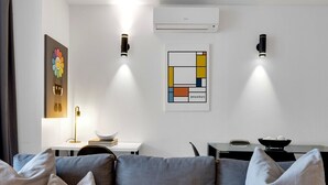 Un salon moderne avec un canapé gris, deux lampes fixées au mur, un appareil de climatisation et une œuvre d’art abstraite encadrée.
