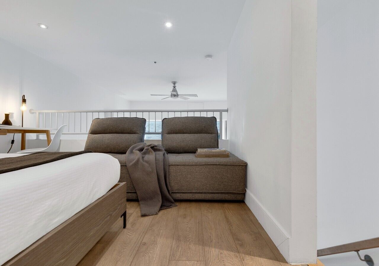 A modern bedroom with a bed, two armchairs, a small table, and a ceiling fan.