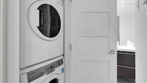 Vue de face d’une buanderie équipée d’un lave-linge et d’un sèche-linge à chargement frontal, d’une porte et d’un plan de travail avec un évier et un miroir.