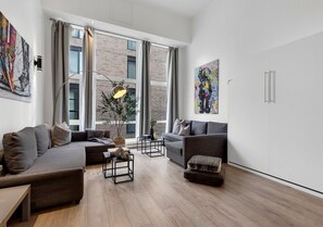 A modern living room with a sectional sofa, a smaller sofa, a coffee table, and a side table. There are two windows with curtains, a large painting on the wall, and a potted plant.
