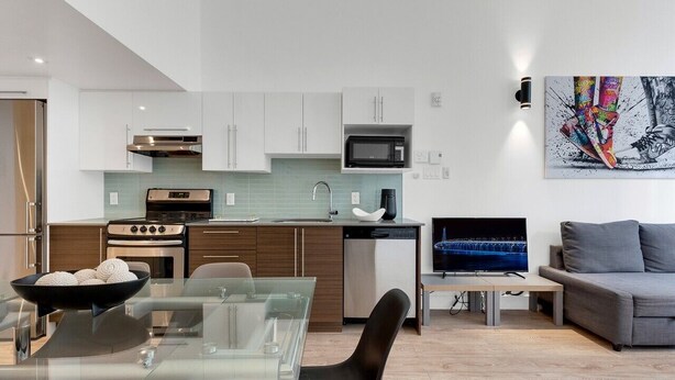 Eine moderne Küche mit weißen Schränken, einer Mikrowelle und einem Geschirrspüler. Es gibt einen Essbereich mit einem Glastisch und Stühlen, und ein graues Sofa im Wohnzimmer.