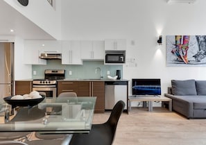 Une cuisine moderne avec des meubles blancs, un four à micro-ondes et un lave-vaisselle. On y trouve un coin repas avec une table et des chaises en verre, ainsi qu’un canapé gris dans le salon.