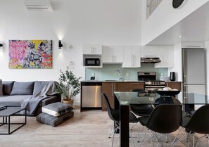 Una cocina moderna con armarios blancos, un refrigerador de acero inoxidable y una zona de comedor con mesa de vidrio y sillas negras.