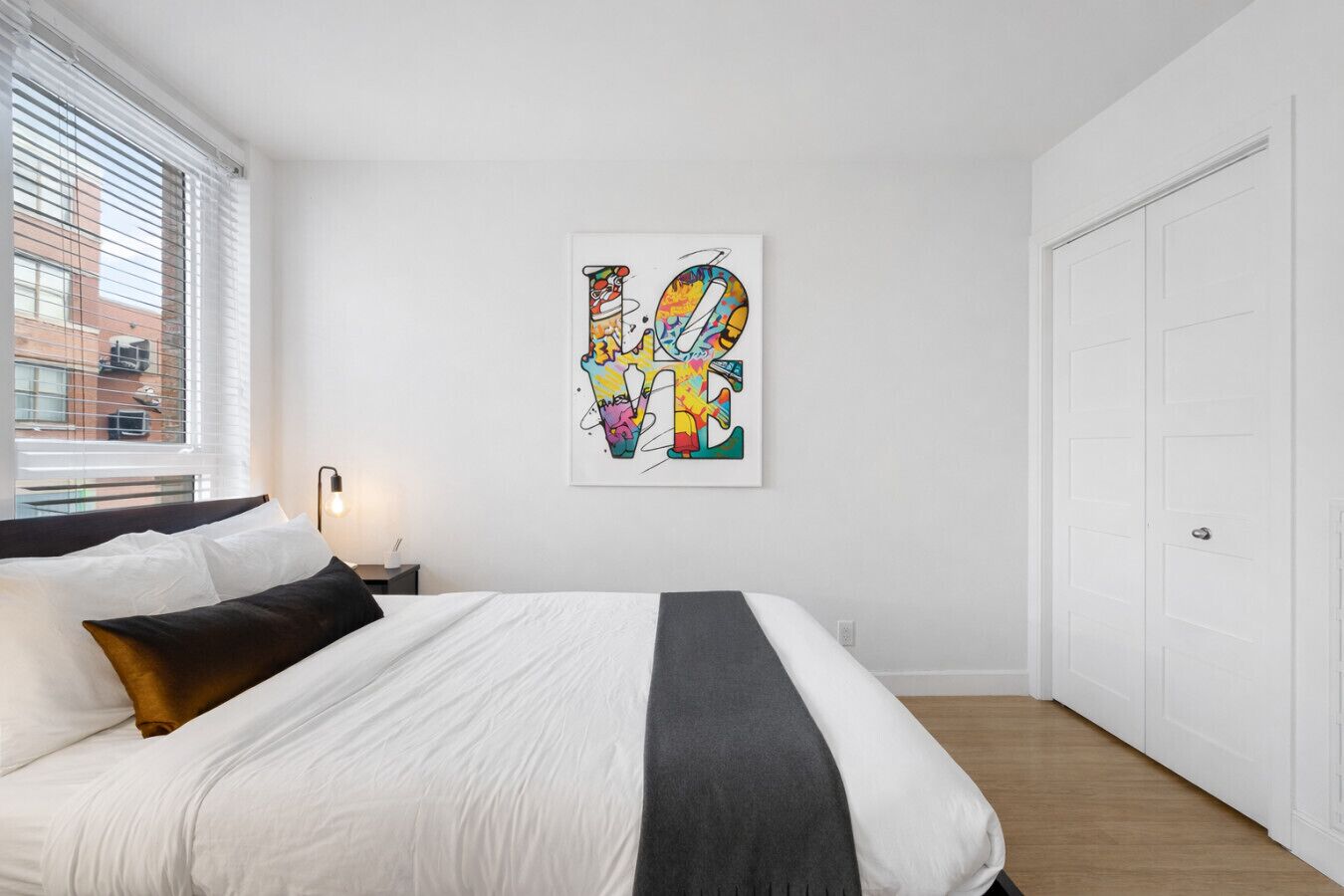 A bedroom with a bed, a nightstand, a lamp, a window with blinds, and a door.