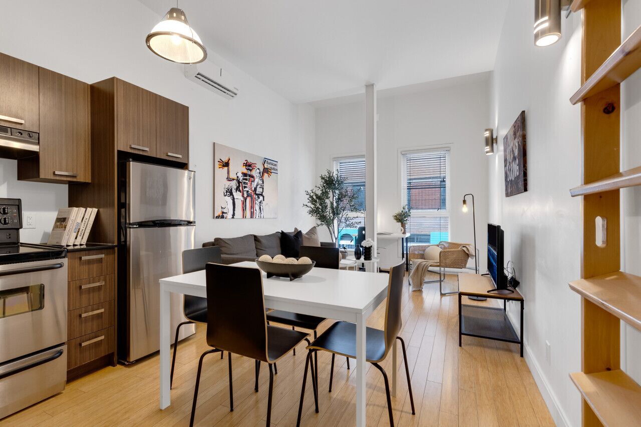 A modern kitchen with stainless steel appliances, a dining area with a white table and black chairs, and a living space with a sofa and a TV.