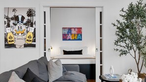Ein modernes Wohnzimmer mit einem grauen Sofa, einem kleinen Beistelltisch mit einer Vase und Büchern, einem großen Gemälde eines Comic-Charakters und einem farbenfrohen abstrakten Kunstwerk an der Wand.