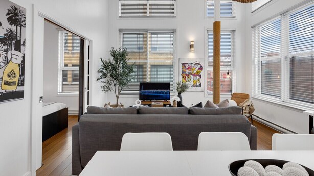 Ein modernes Wohnzimmer mit einem grauen Sofa, einem Fernseher und einem großen Fenster mit Jalousien.