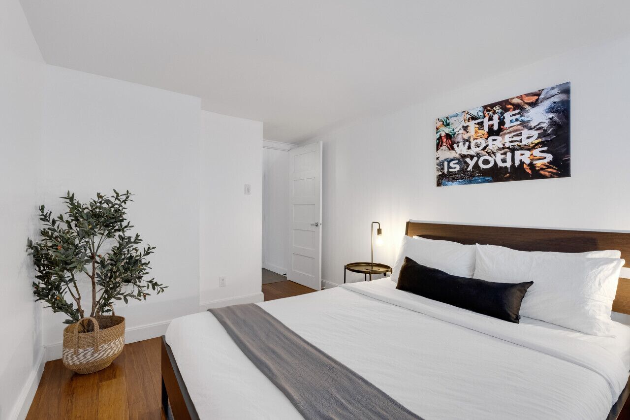 A neatly made bed with white and dark pillows, a small bedside table with a lamp, and a wall-mounted artwork.