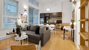 A modern living room with a grey sofa, white coffee table, and a dining area with a white table and chairs. A kitchen with dark cabinets and stainless steel appliances is visible in the background. - LiV MTL Main Plateau Urban Suites Near Downtown Montreal (Montréal)