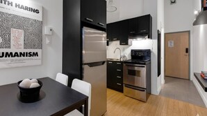 A modern kitchen with a black refrigerator, stove, and oven, a dining table with a bowl of fruit, and a wall with two framed posters.