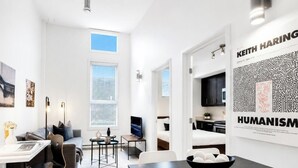 A modern hotel room with a large window, a flat-screen TV, a dining area, and a poster on the wall.