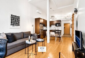 A modern living room with a sofa, coffee table, and TV. There's a kitchen area with a dining table and chairs.