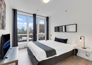 A modern hotel room with a large bed, a desk, a TV, and a view of the city through large windows. - Spacious 3BR Penthouse Rooftop • Sleeps 12 (Montréal)