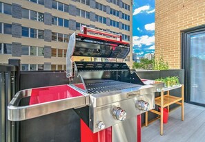 A stainless steel outdoor grill with a red and black design, a wooden chair, and a potted plant on a balcony with a brick building in the background.