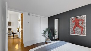 A bedroom with a bed, a dark wall with a red abstract painting, a white door, and a hallway leading to a kitchen.