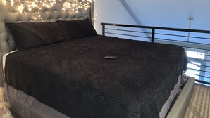 Un dormitorio con cabecera capitonada, ropa de cama oscura y un control remoto sobre la cama.