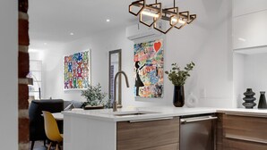 Una cocina moderna con encimera blanca, armarios de madera y lavavajillas de acero inoxidable. Hay dos cuadros abstractos y coloridos en la pared, una planta en maceta y una lámpara colgante.
