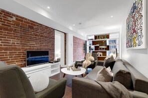A modern living room with a brick wall, a flat-screen TV, a sofa, a coffee table, and a bookshelf.