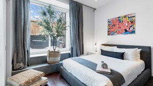 A modern bedroom with a large bed, a window with curtains, a plant, and a colorful artwork on the wall. - LiV MTL Soho Spacious Rooftop Suites for Groups Near Downtown Montreal (Montréal)