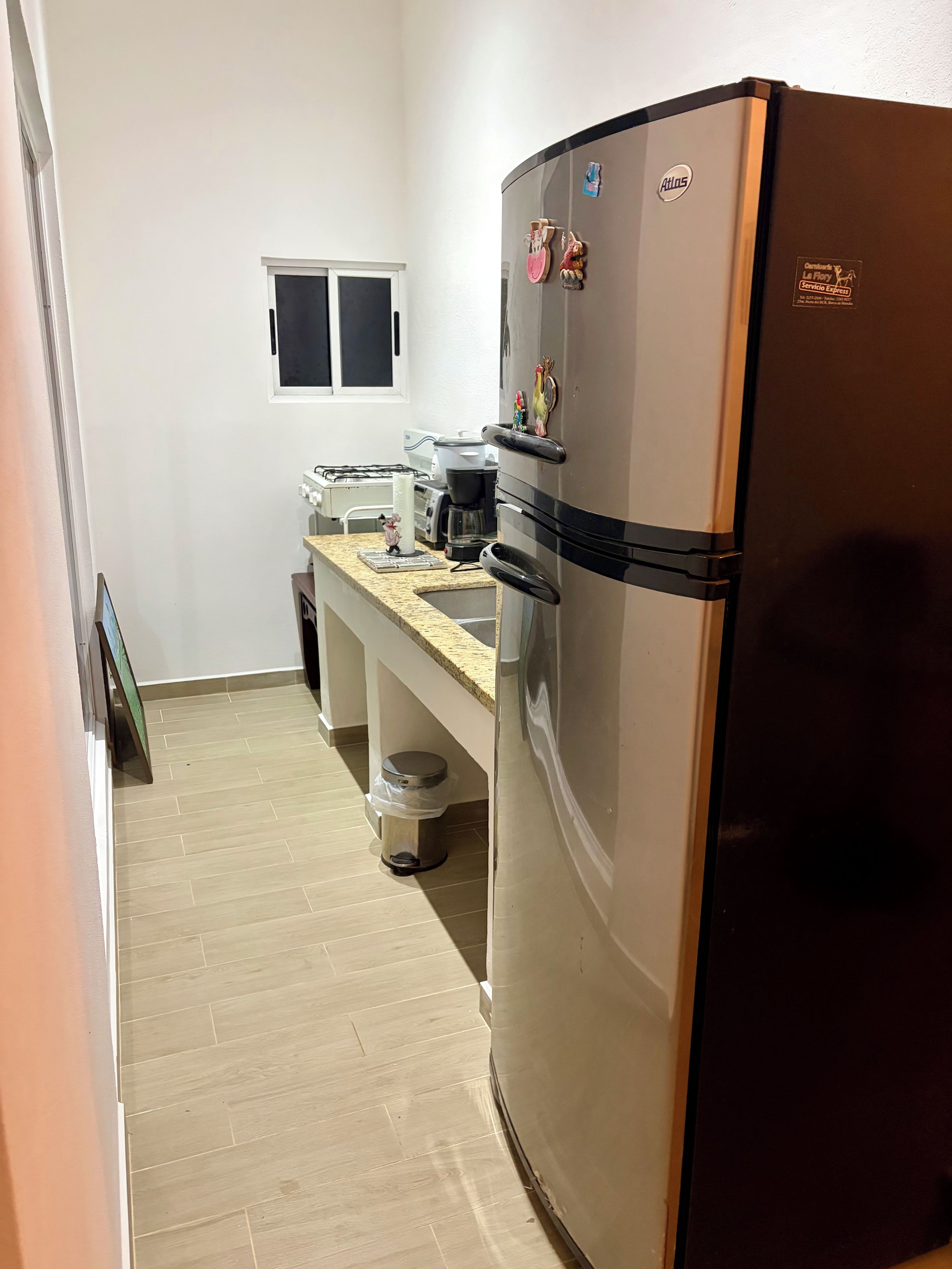 Cocina compacta con refrigerador de acero inoxidable, una encimera con cafetera y un cesto de basura.