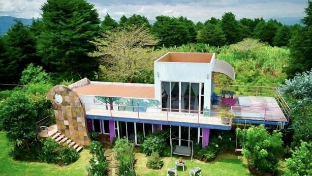 Una casa moderna con un invernadero de vidrio, una escalera de piedra y un pequeño jardín con mobiliario exterior.