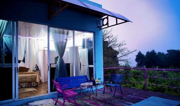 Una casa moderna con un vidrio que da a un dormitorio, un balcón con mesa y sillas, y un toldo azul.