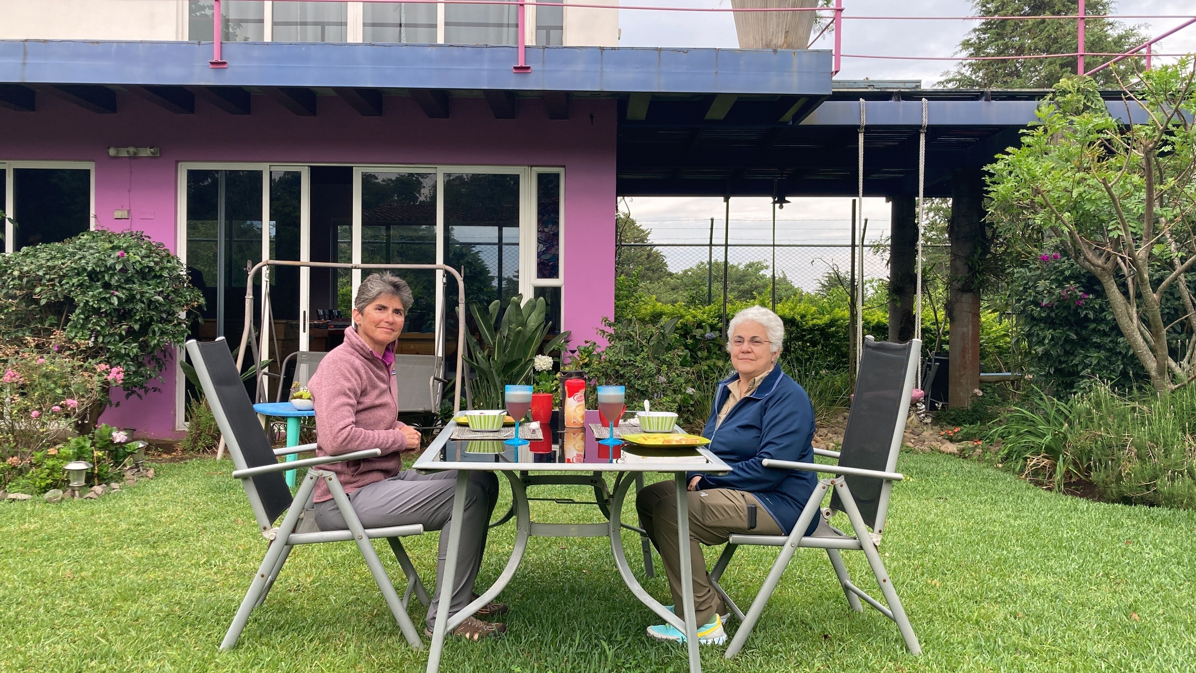 Two people seated at an outdoor table with a garden view, a purple building in the background, and a swing set.