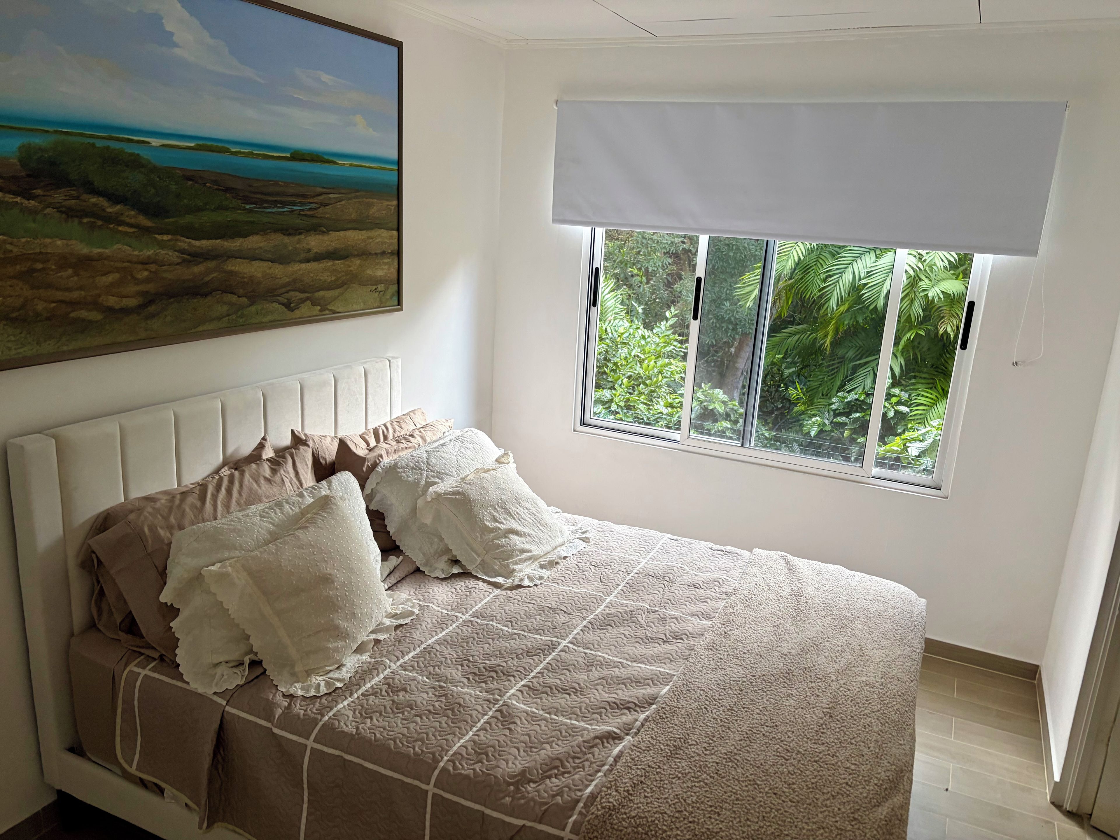 A bedroom with a bed, a window with a view of greenery, a painting on the wall, and a ceiling with a textured finish.
