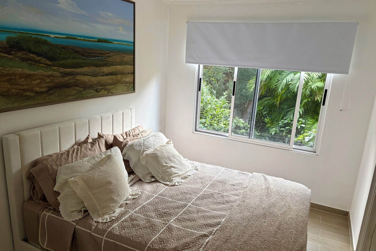 A bedroom with a bed, a window with a view of greenery, a painting on the wall, and a ceiling with a textured finish.