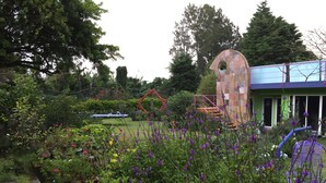 Un jardín con diversas plantas y flores, una estructura de piedra singular y un edificio con techo azul.