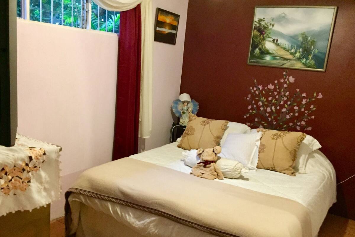 A neatly made bed with a teddy bear, a vase of flowers, and a painting on the wall.